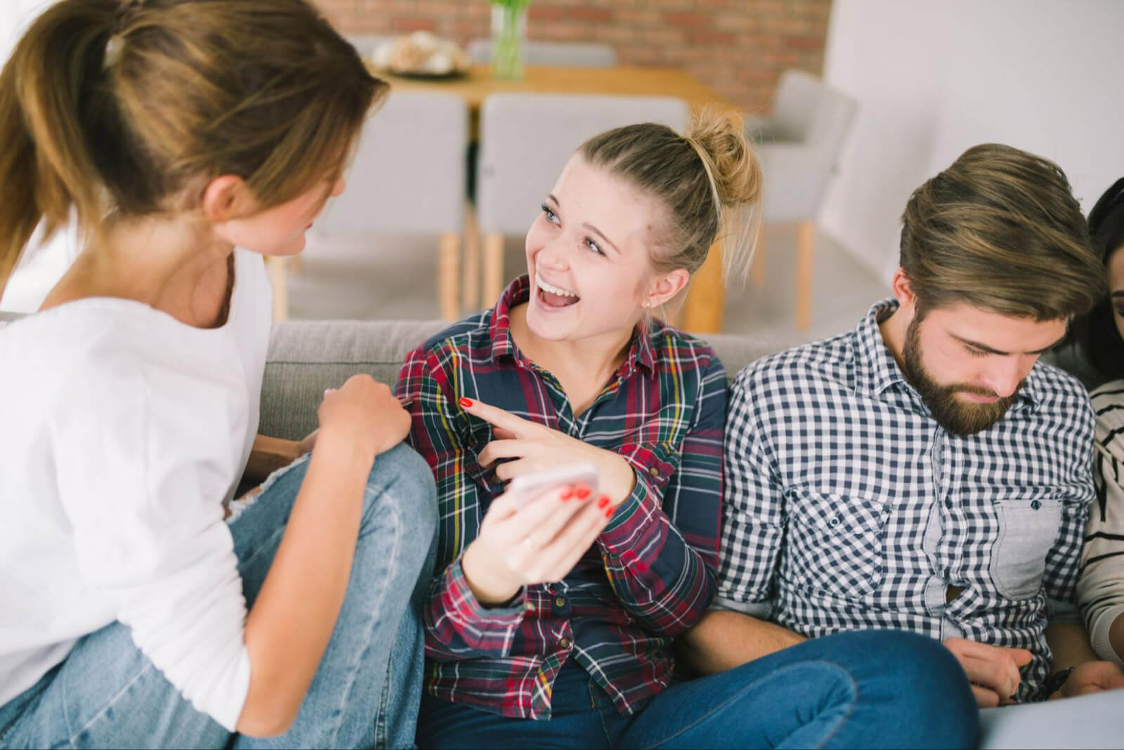 A group of teens sit together on a couch, talking and reacting to something on a phone in a casual home environment.