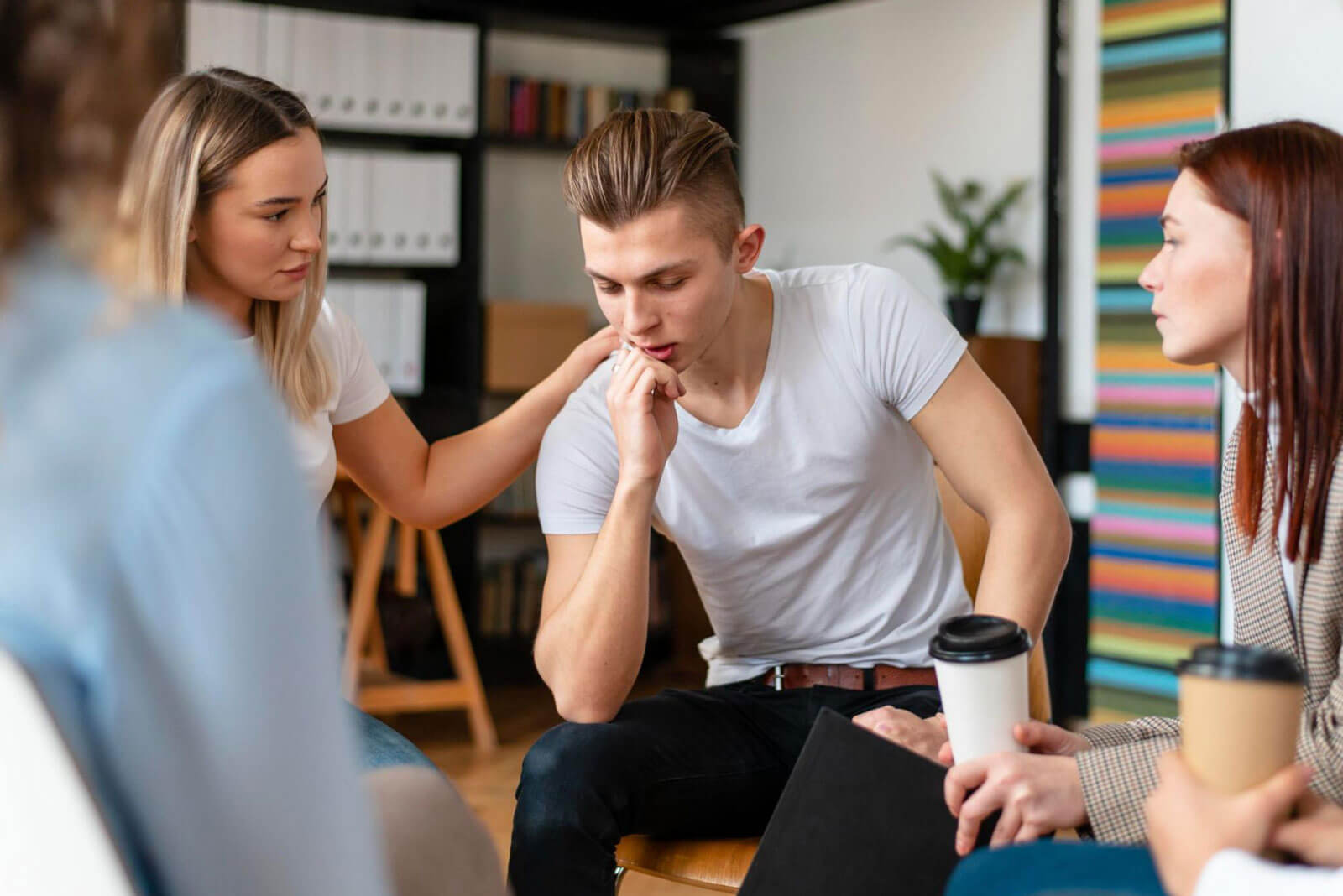 A young man sitting in a support group with others, looking distressed while a woman places a reassuring hand on his shoulder.
