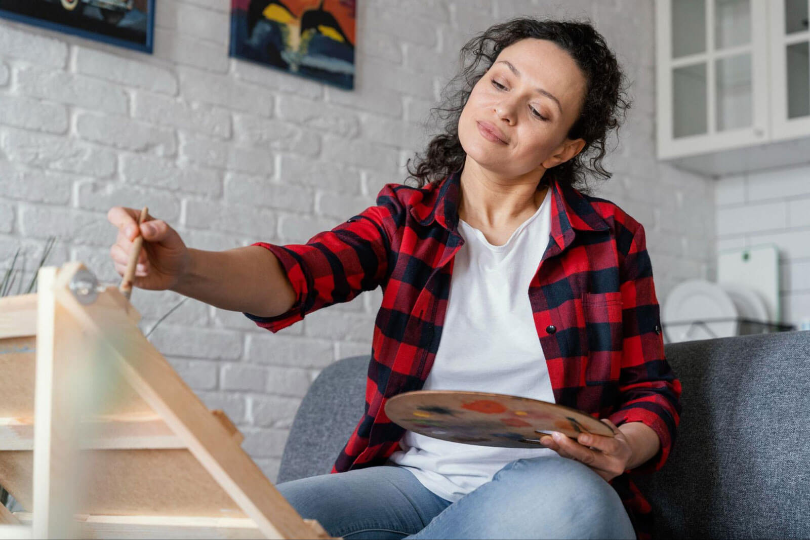 A woman sitting on a couch paints on an easel while holding a paint palette.